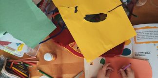A child drawing on orange paper at a wooden table with craft supplies