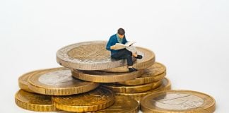 Miniature person sitting on stack of coins reading newspaper