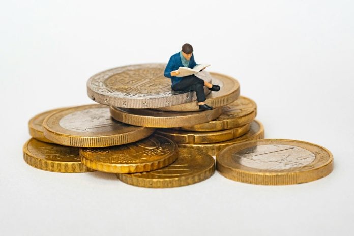 Miniature person sitting on stack of coins reading newspaper